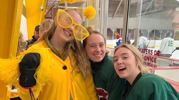 Tres estudiantes de la Rideau District High School posan sonriendo en una arena deportiva con equipos deportivos de fondo.