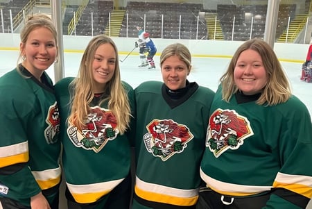 Cuatro estudiantes de la Rideau District High School llevan camisetas de hockey verdes y están juntas en la superficie de hielo de la sala de hockey.