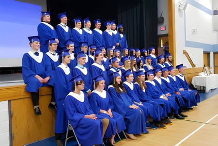 Un gran grupo de graduadas y graduados de la Ridgetown District High School está en el escenario con togas azules durante la ceremonia de graduación.