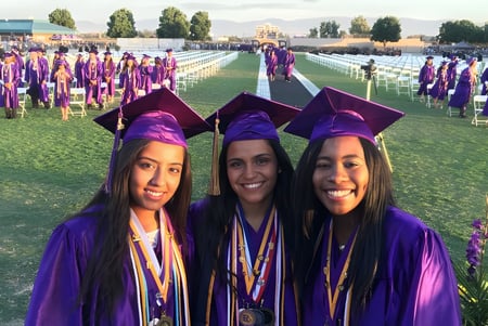 Tres graduadas de la Ridgeview High School en vestimenta de graduación morada están frente a un gran grupo de graduados en un campo de hierba.
