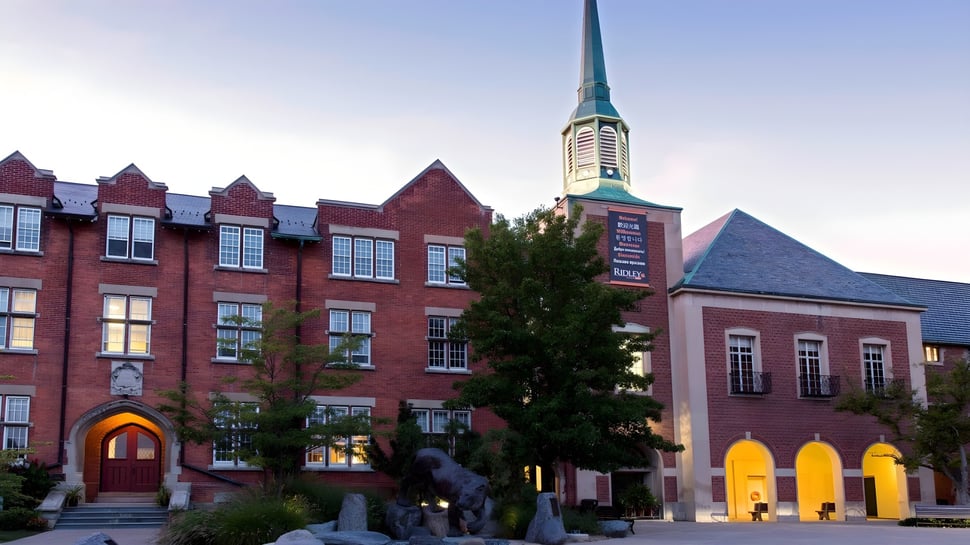El edificio histórico de ladrillo con alto torre en el terreno del Ridley College está rodeado de árboles y otros edificios.