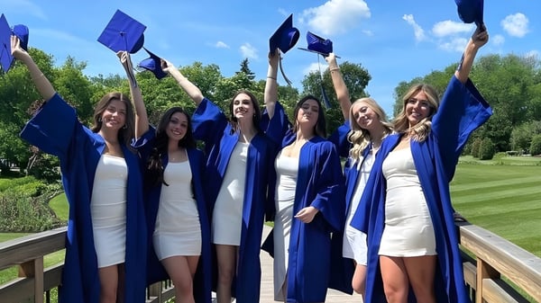Un grupo de graduadas de la River East Collegiate celebra al aire libre con los brazos en alto y diplomas.