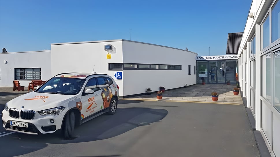 Un BMW X1 blanco está frente a un gran edificio blanco en el campus de la Rockford Manor School bajo un cielo despejado.