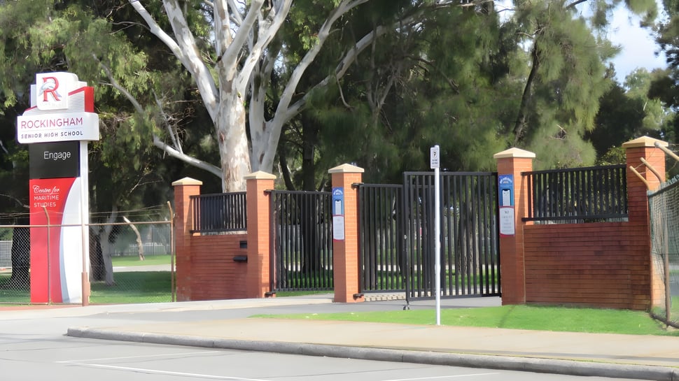El área de entrada de la Rockingham Senior High School con pilares de mampostería y una cerca de metal frente a una superficie de césped con árboles.