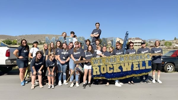 Un grupo de jóvenes posa frente a un banner de la Rockwell Charter School con un paisaje montañoso de fondo.