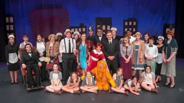 Un gran grupo de estudiantes de Rocky Bayou Christian High School posan en disfraces y ropa formal en un escenario con fondo azul.