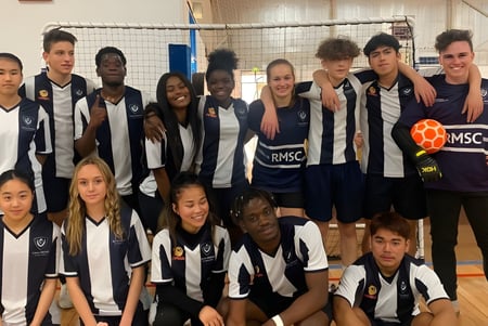 Un grupo de estudiantes del Roma Mitchell Secondary College posan juntos en un gimnasio con una red de voleibol al fondo.