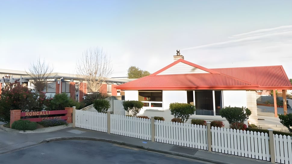 El Roncalli College muestra una casa con techo rojo y una cerca blanca de listones en primer plano y árboles al fondo.