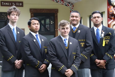 Un grupo de estudiantes en ropa formal está frente al edificio del Rongotai College con el letrero Tango to Kio.