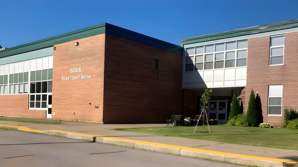 El edificio de ladrillo de la Rose-Des-Vents School con techo verde y grandes ventanas se encuentra en un área pavimentada bajo un cielo azul.