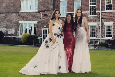 Tres alumnas de la Rossall School en vestidos festivos están frente a un edificio de ladrillo con grandes ventanas en un área de césped.
