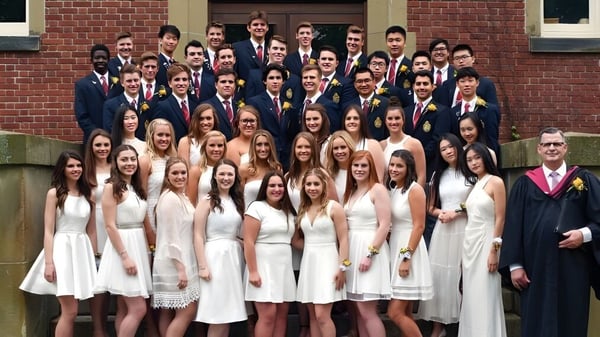 Un gran grupo de estudiantes está de pie en ropa formal frente a un edificio de ladrillo de la Rothesay Netherwood School.