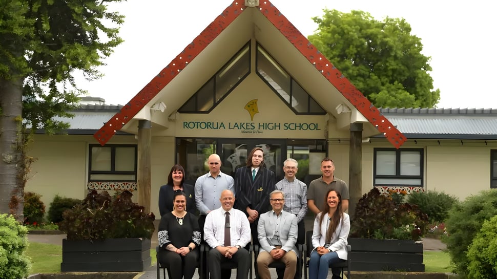 Empleados de la Rotorua Lakes High School están frente al edificio escolar con un distintivo techo triangular en un entorno verde.