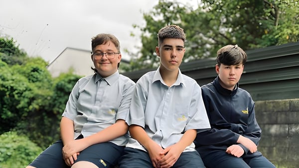 Tres estudiantes en uniformes escolares están sentados juntos al aire libre en el terreno de la Rotorua Lakes High School frente a un fondo verde.