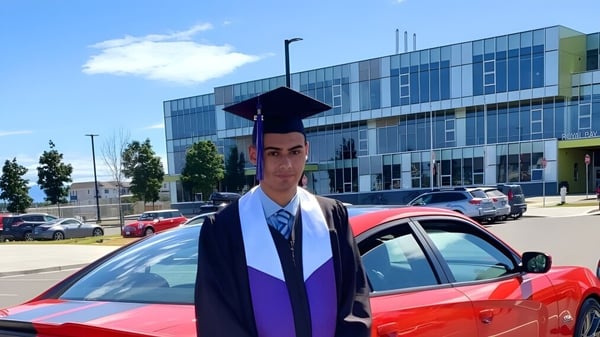 Un estudiante de la Royal Bay Secondary School está en toga de graduación frente a un auto rojo ante un edificio moderno.