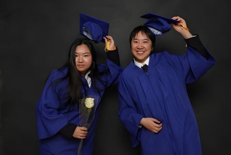 Dos estudiantes de la Royal Crown School celebran su graduación en togas azules frente a un fondo negro.