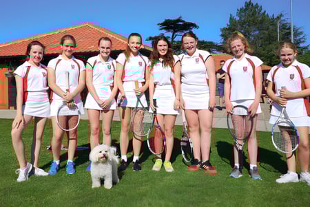 Un grupo de alumnas y alumnos de la Royal Hospital School vestidos de tenis está con un perro en un césped frente a un edificio con techo de tejas rojas.