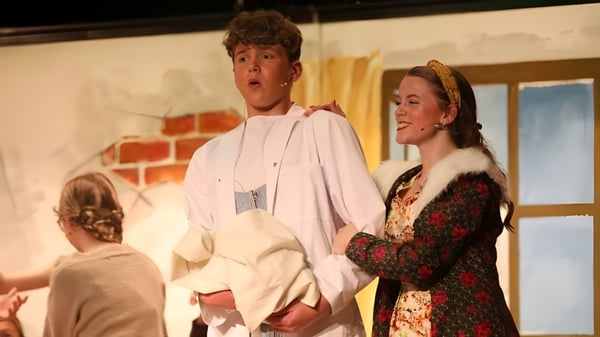 Dos personas están en el escenario de la Royal School Armagh frente a una pared de ladrillos con atrezzo de escenario en el fondo.