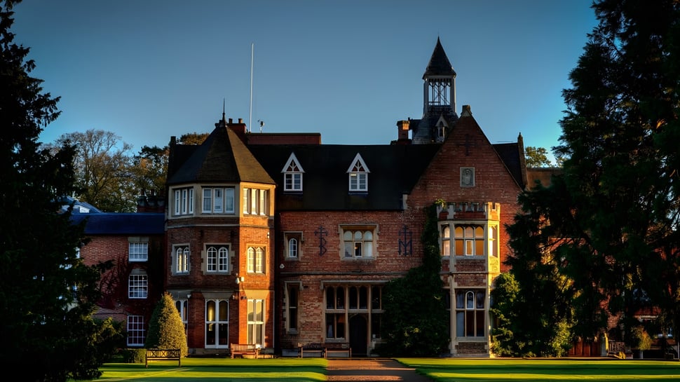 El histórico edificio de ladrillo de la Rugby School está rodeado de césped y árboles y se encuentra bajo un cielo azul claro.