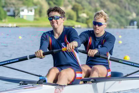 Dos alumnas en uniforme deportivo están sentadas en un bote sobre el agua frente a un paisaje natural en el campus del Sacred Heart College.