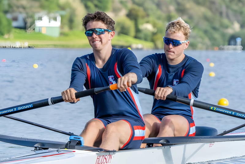 Dos alumnas en uniforme deportivo están sentadas en un bote sobre el agua frente a un paisaje natural en el campus del Sacred Heart College.
