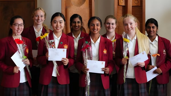 Estudiantes del Sacred Heart Girls' College Hamilton llevan togas rojas de graduación y sostienen certificados en una sala con paneles de madera.