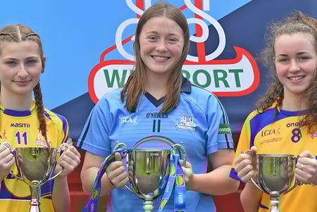Tres alumnas de la Sacred Heart School en uniformes deportivos sostienen trofeos frente a un logo de Waterford.