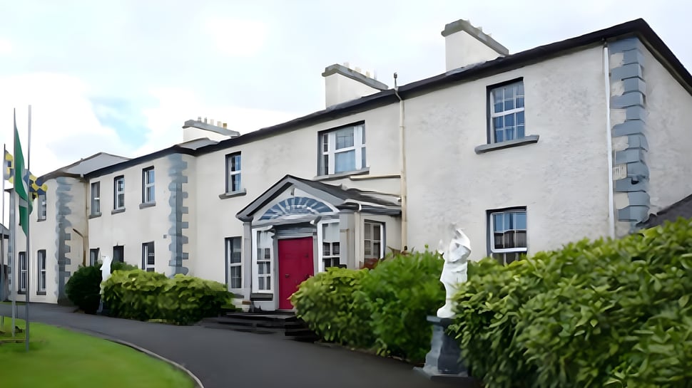 El edificio de varios pisos de la Sacred Heart School con puerta roja está rodeado de árboles verdes y una superficie de césped.