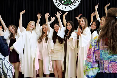 Un grupo de jóvenes alumnas de la Sacred Heart School of Halifax está en túnicas blancas en un escenario con los brazos levantados frente a un gran logo.