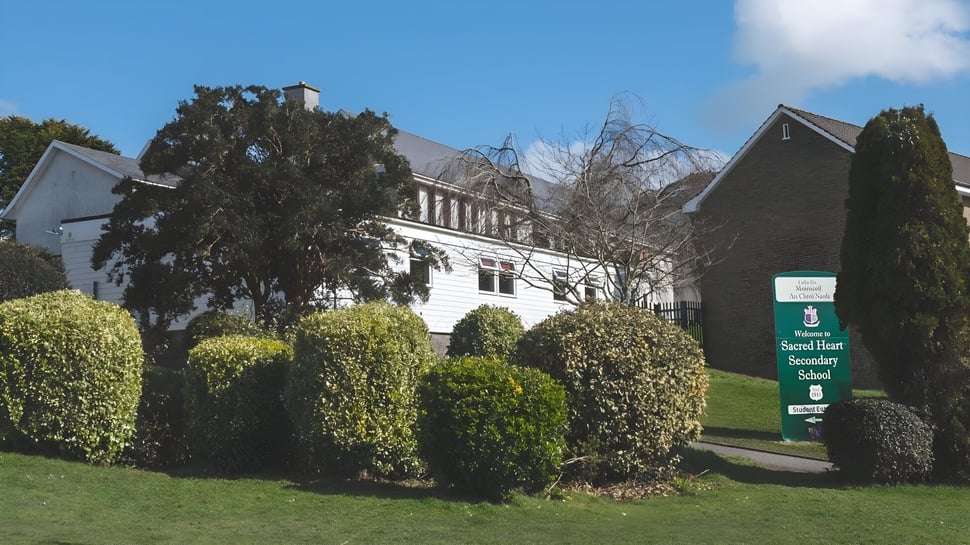 El edificio histórico de la Sacred Heart Secondary School Clonakilty está rodeado de setos y árboles bien cuidados bajo un cielo azul claro.