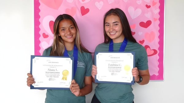 Dos estudiantes de la Sacred Hearts Academy sostienen sus certificados en alto frente a un fondo rosa con motivos de corazones.