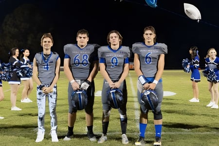 Un grupo de cheerleaders de la Saint Francis Catholic Academy está de pie por la noche en el campo de fútbol bajo una brillante iluminación.