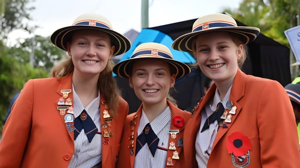 Tres estudiantes en uniformes naranjas están sonriendo frente a árboles en el campus del Saint Stephen’s College.
