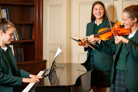 Tres alumnas de la Salerno Secondary School tocan juntas el violín y el piano en una sala de música con estanterías.