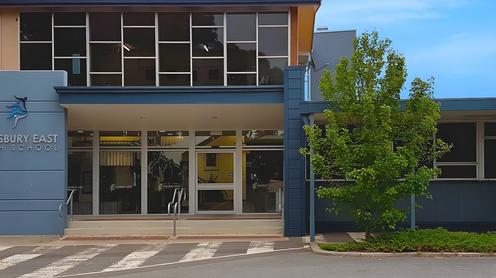 El moderno edificio de varios pisos de la Salisbury East High School con grandes ventanas y un árbol en primer plano bajo un cielo azul.