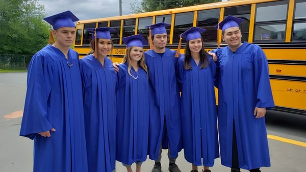 Un grupo de graduados en togas azules se encuentra frente a un autobús escolar amarillo en el terreno de la Salmo Secondary School.