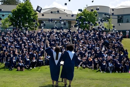 Un grupo de graduados de la Salmon Arm Secondary en togas negras está frente a un edificio en un campo de césped celebrando su graduación.