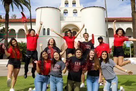 Un grupo de jóvenes estudiantes con camisetas rojas posan frente a un gran edificio blanco con reloj de torre en el campus de la San Diego State University.