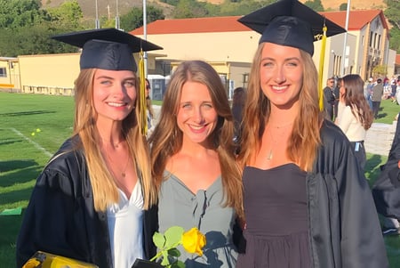 Tres alumnas en gorros y togas de graduación están juntas frente a un césped en el campus del San Luis Coastal Unified School District.