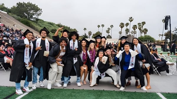 Un grupo de graduados del Santa Barbara City College posan al aire libre frente a árboles y un campo.