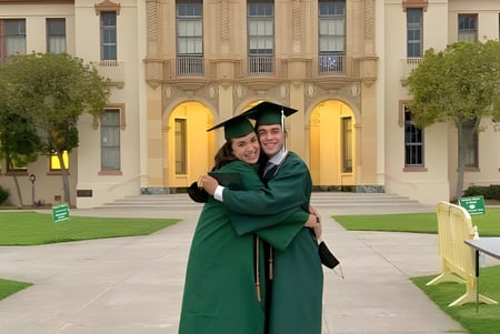 Dos personas con birretes y togas de graduación se abrazan frente al edificio histórico de la Santa Barbara High School.