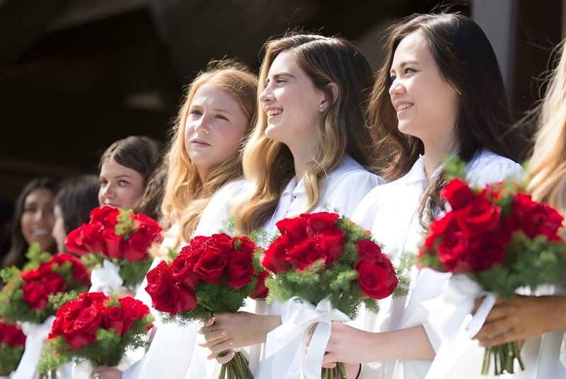 Un grupo de jóvenes mujeres en vestidos blancos sostiene rosas rojas frente a un fondo oscuro en el terreno de la Santa Catalina School.