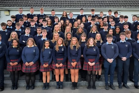 Un grupo de alumnas y alumnos en uniforme escolar está frente a un edificio con techo de tejas en el campus de Scoil Mhuire (Strokestown).