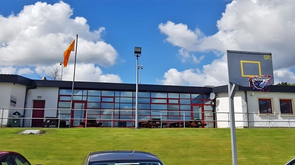 El moderno edificio de la Scoil Ruáin con un aro de baloncesto se encuentra en un prado verde bajo un cielo azul.