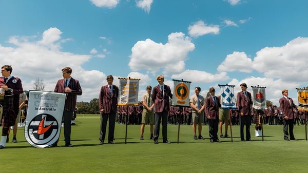 Un grupo de estudiantes del Scotch College está en un campo de hierba con ropa tradicional bajo un cielo azul con nubes blancas.
