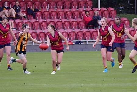 Un grupo de jóvenes atletas en uniformes rojos y amarillos corre en un campo en Scotch Oakburn College frente a espectadores.