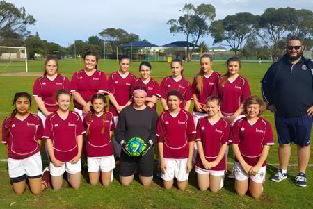 Un grupo de jóvenes futbolistas en camisetas rojas posan con un entrenador en un campo de césped en el Seaford Secondary College.