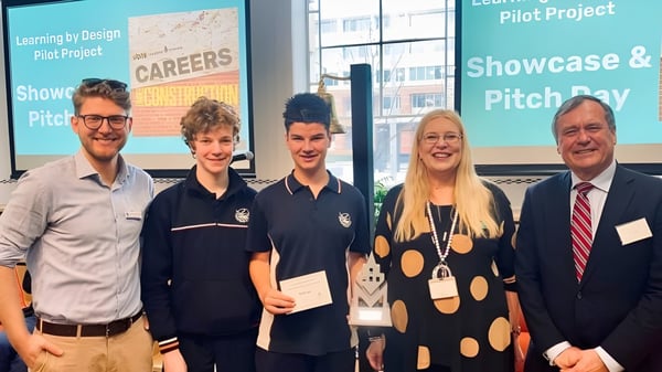 Un grupo de estudiantes y un profesor están frente a un panel informativo sobre el proyecto piloto Learning by Design en el campus de la Seaton High School.