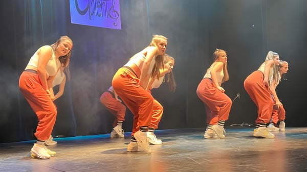 Cuatro bailarinas y bailarines en trajes naranjas realizan una presentación de baile en el escenario del Séminaire de Chicoutimi con un fondo azul-púrpura.