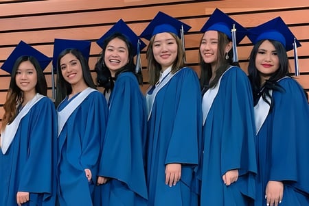 Un grupo de estudiantes en túnicas de graduación azules celebra frente a una pared de madera en el terreno de la Sentinel Secondary School su graduación.